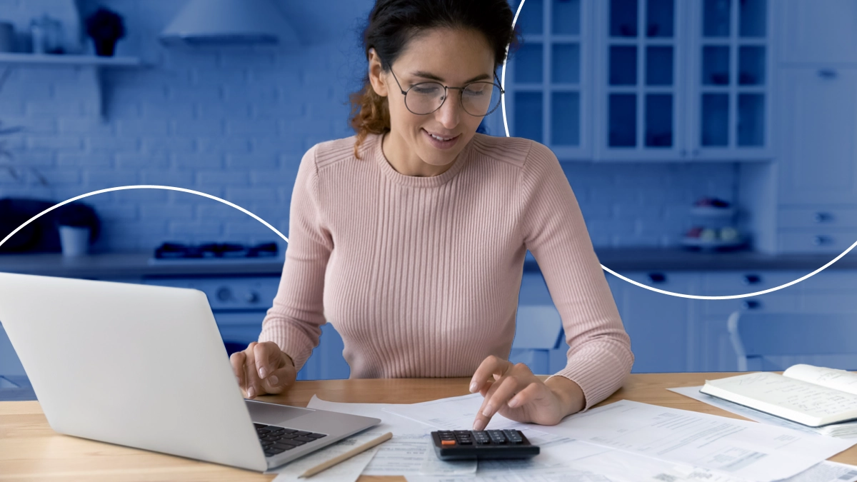 Mulher sorrindo, mexendo e olhando para uma calculadora que está sobre uma mesa com papéis e um notebook