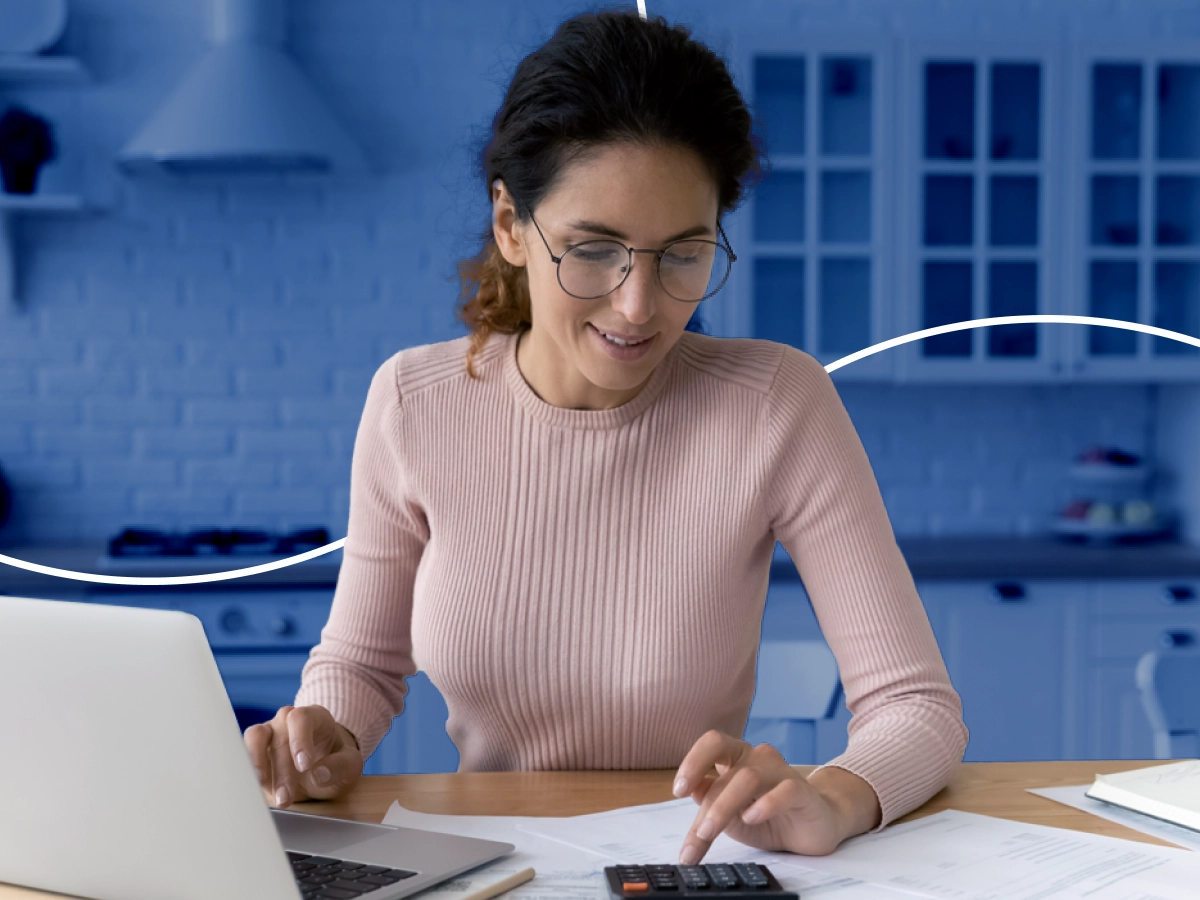 Mulher sorrindo, mexendo e olhando para uma calculadora que está sobre uma mesa com papéis e um notebook